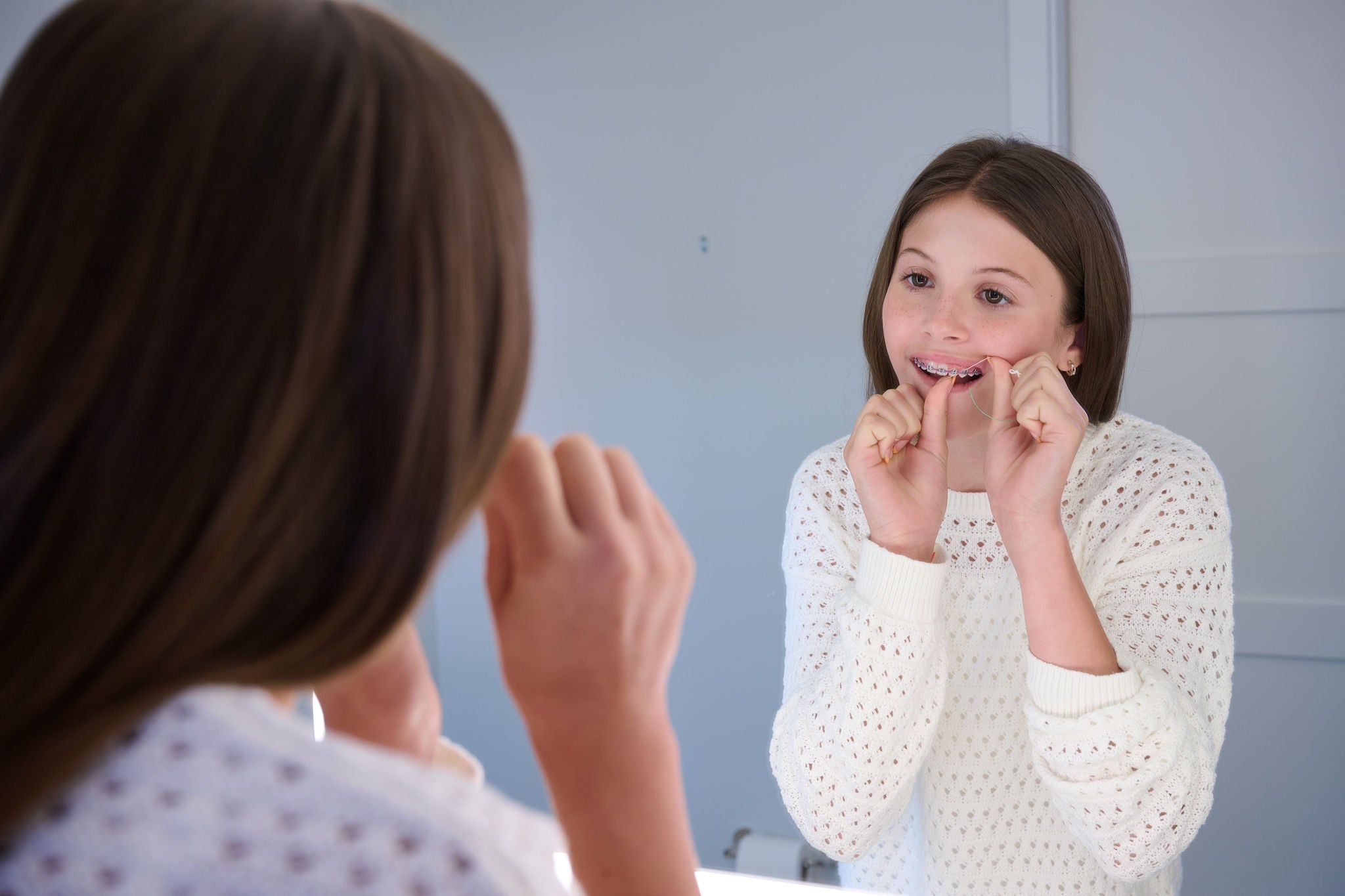 A girl flosses her teeth with string floss in front of a mirror. She wears a white sweater. Image resolution: 2048x1365.