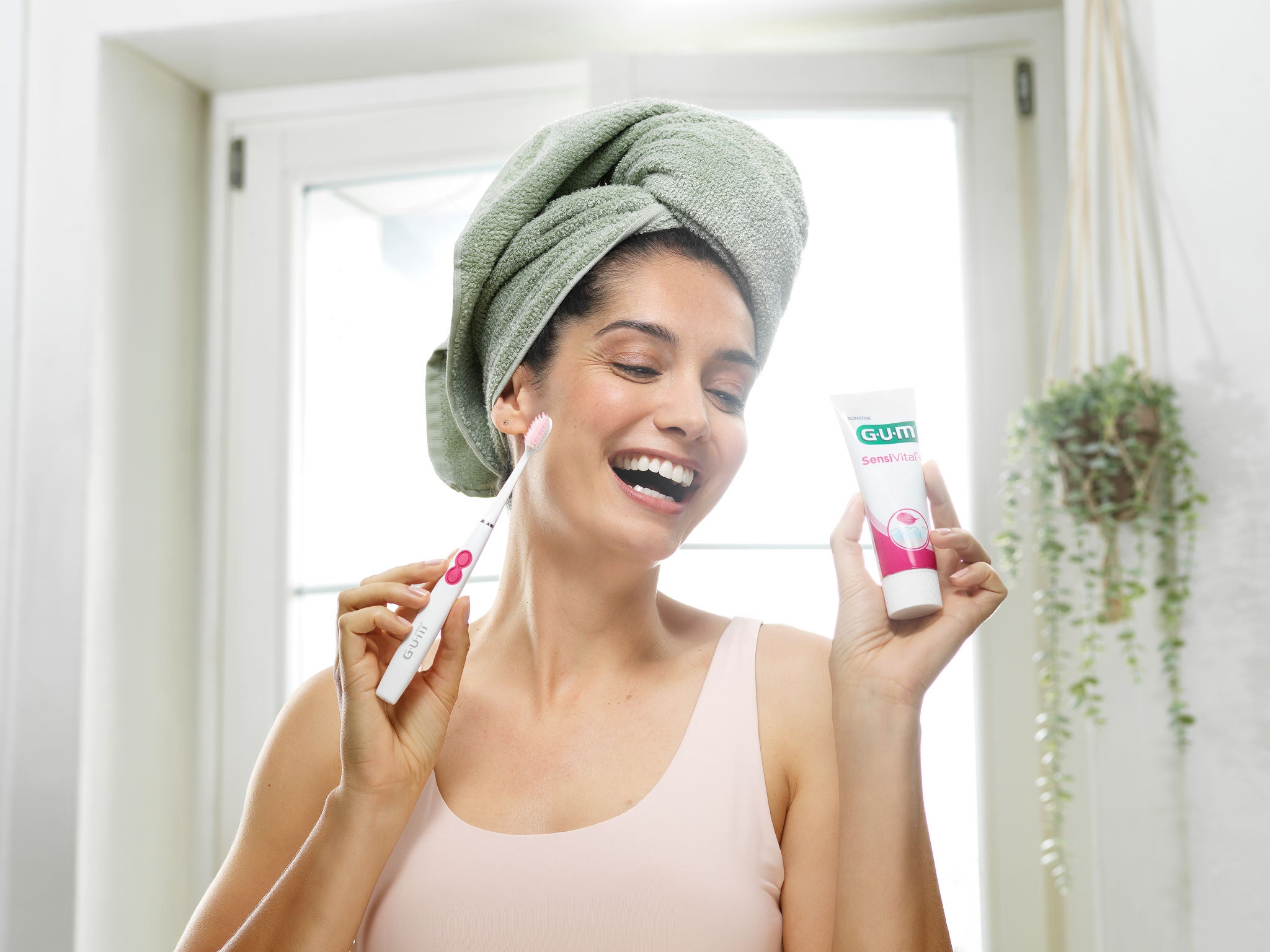 Smiling woman is holding the GUM SONIC SENSITIVE battery toothbrush and the GUM SensiVital toothpaste