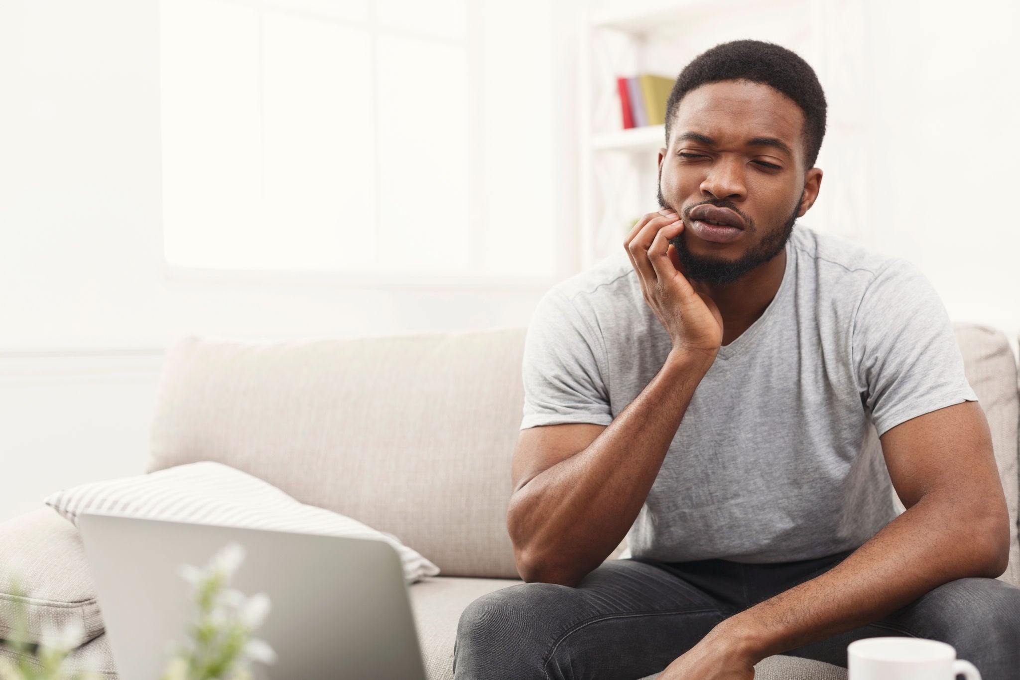 Young african-american businessman suffering from toothache while working on laptop, sitting on couch at home, copy space