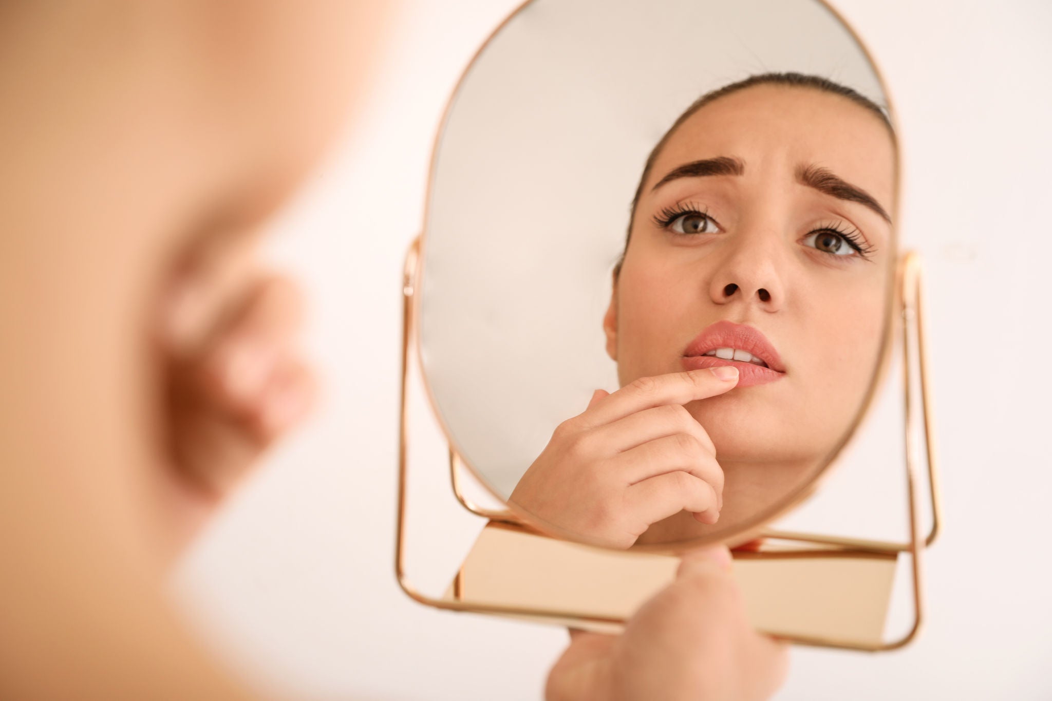 Emotional woman with herpes touching lips in front of mirror against light background