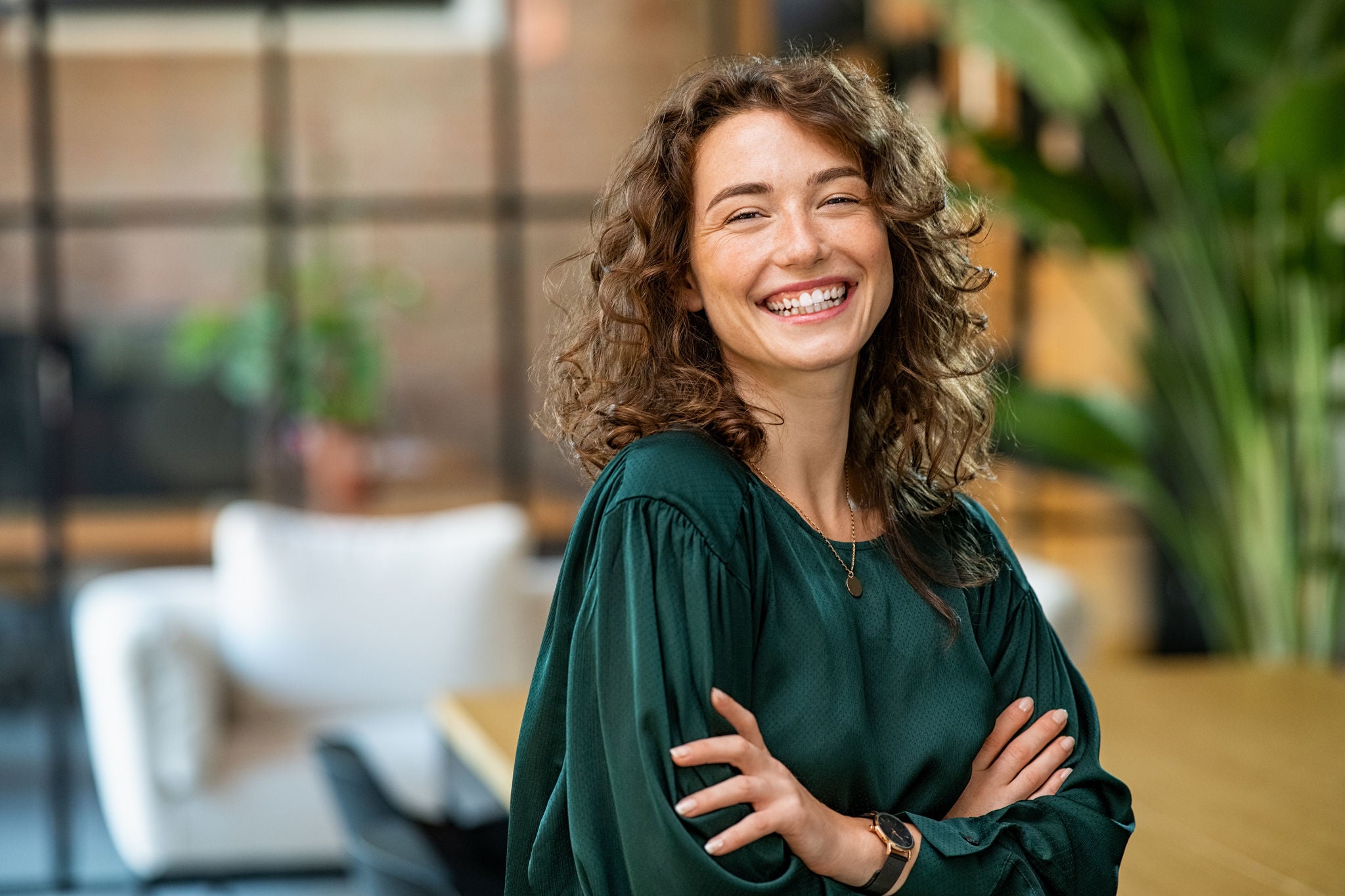 Portrait of young smiling woman looking at camera with crossed arms. Happy girl standing in creative office. Successful businesswoman standing in office with copy space.