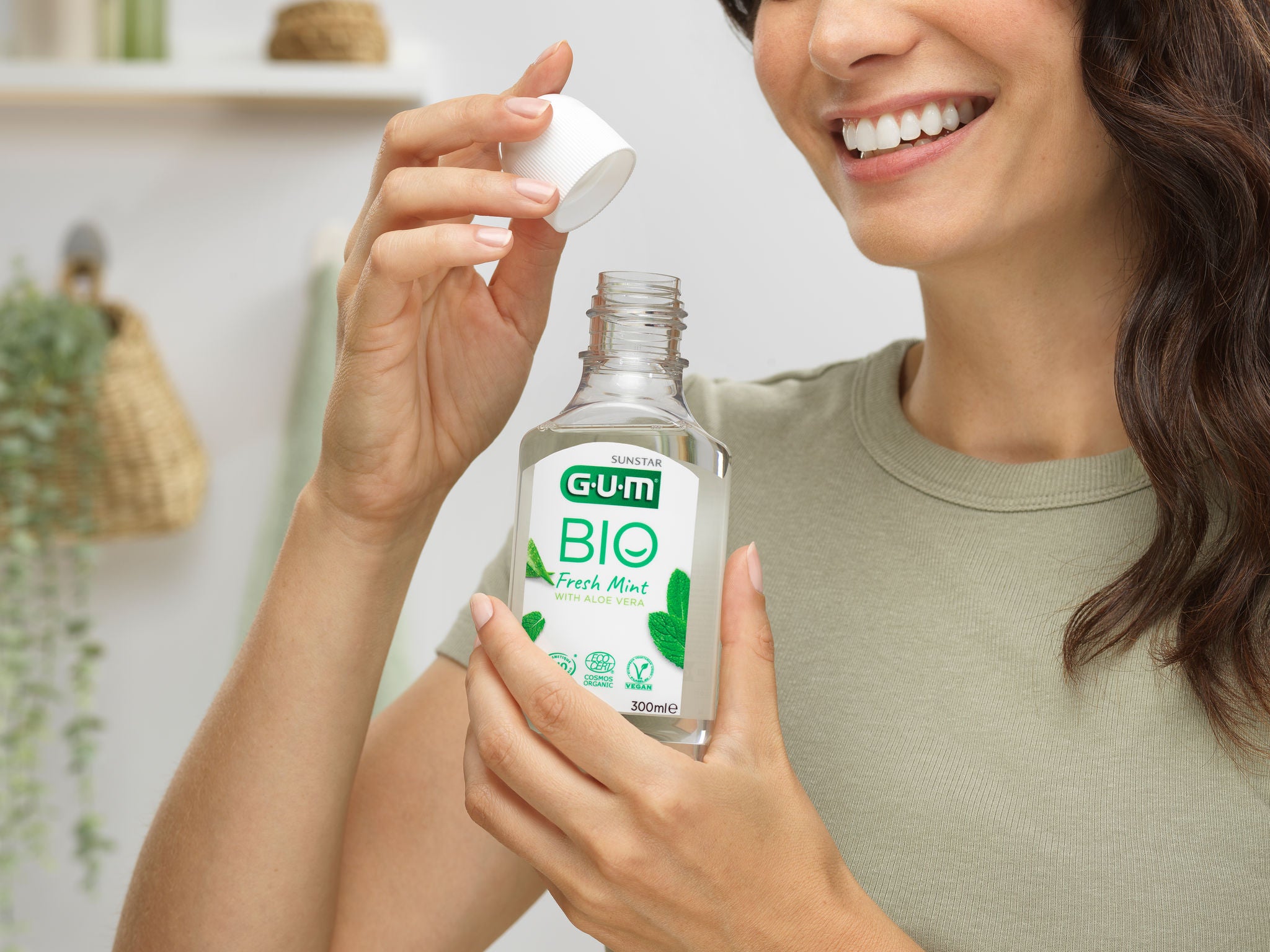 Woman is smiling and is opening a bottle of GUM BIO Mouthwash in a natural environment