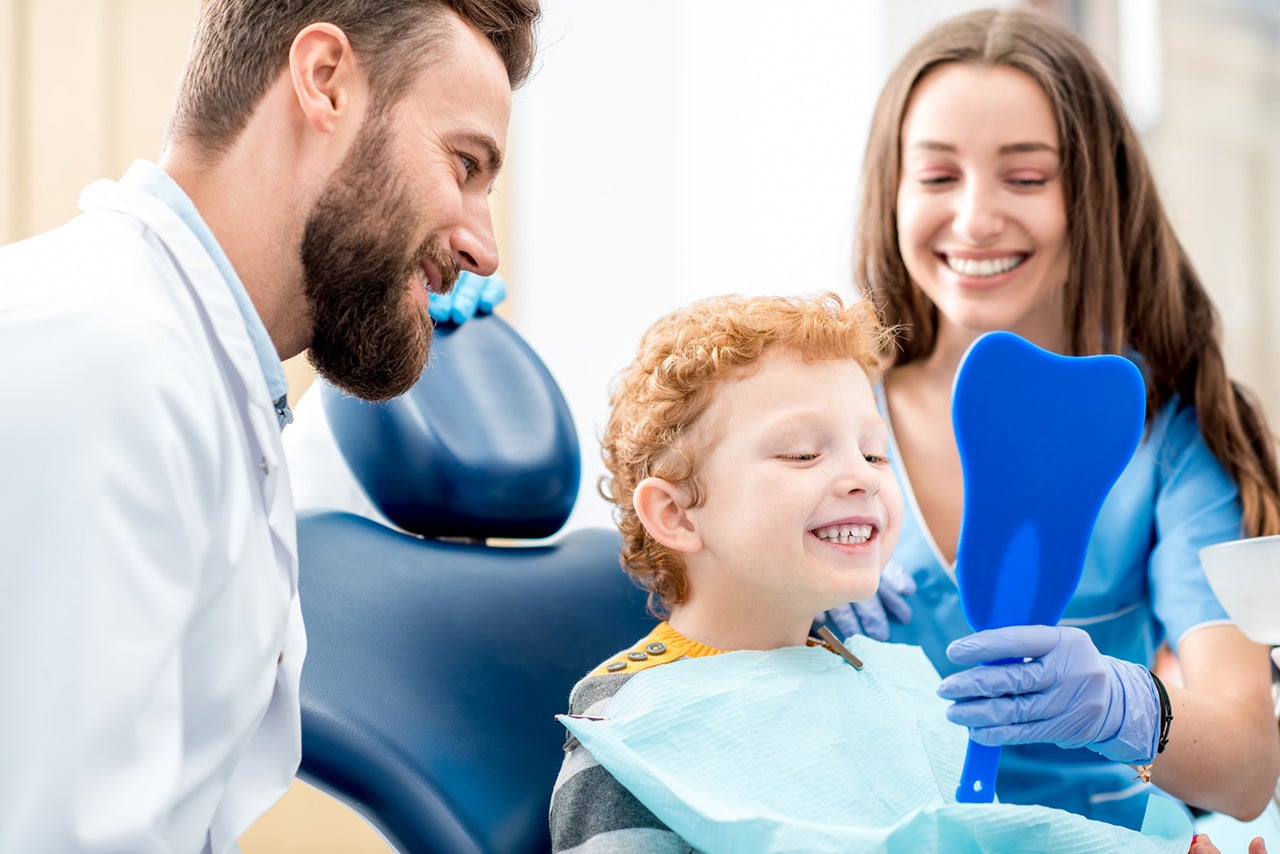 Young boy looking at the mirror with toothy smile sitting on the chair with dentist and assistant at the dental office