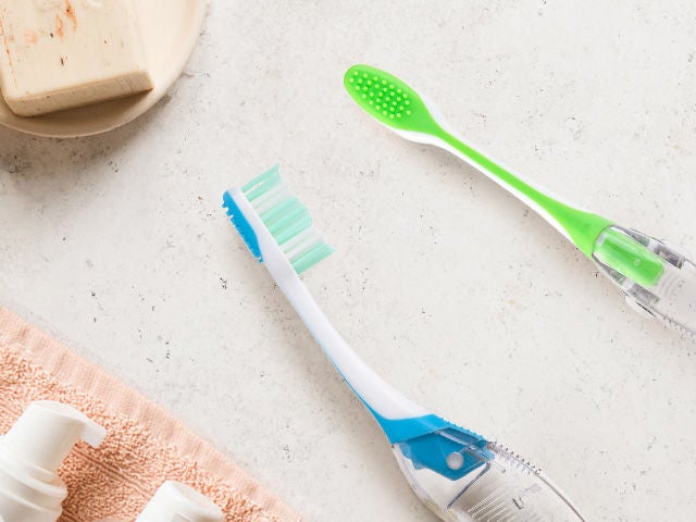 Two GUM TARVEL toothbrushes in blue and green colours shown from the side and back, placed in a natural environment