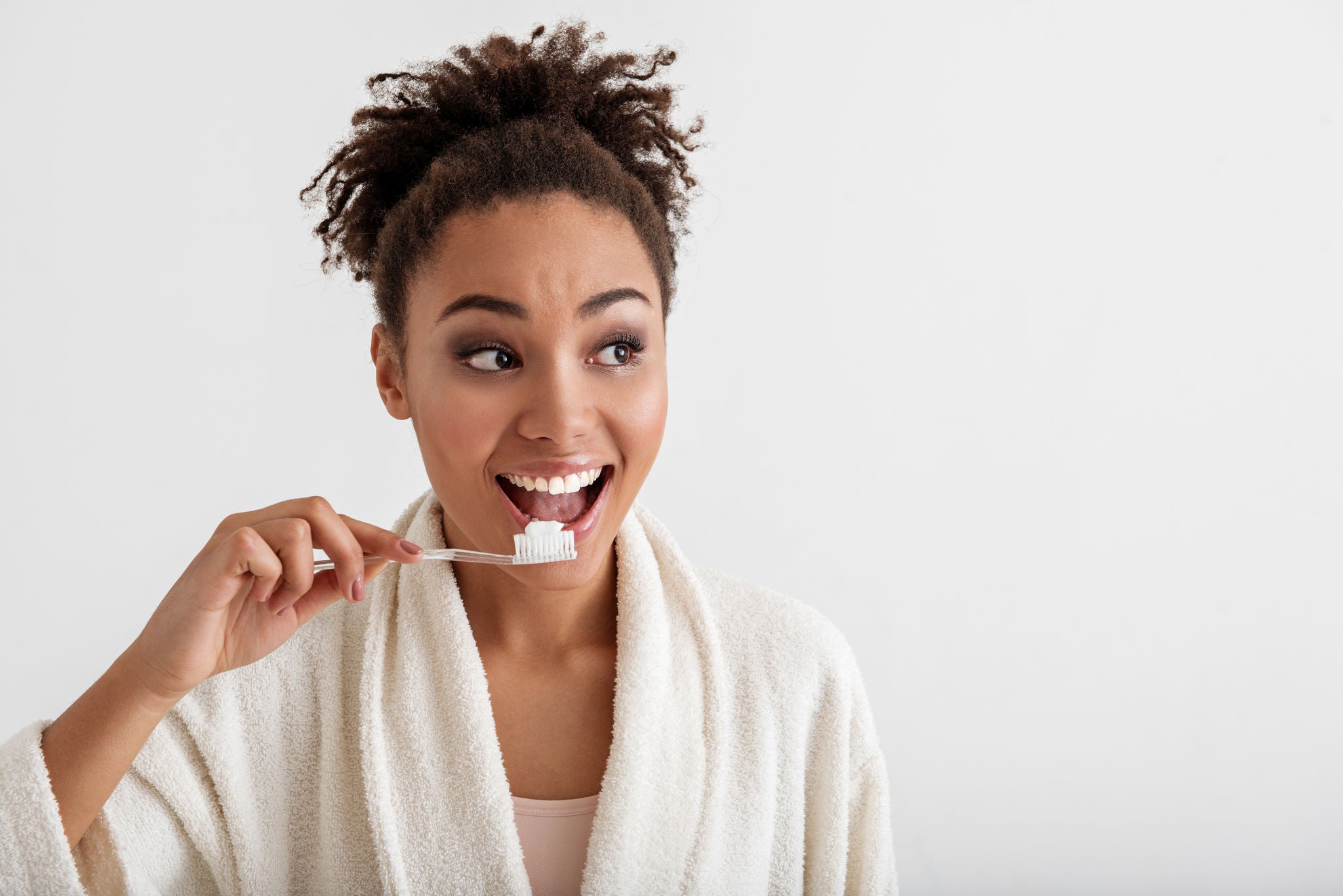 Portrait of smiling girl brushing teeth. She is wearing housecoat and looking aside with happiness. Copy space in right side. Isolated on background