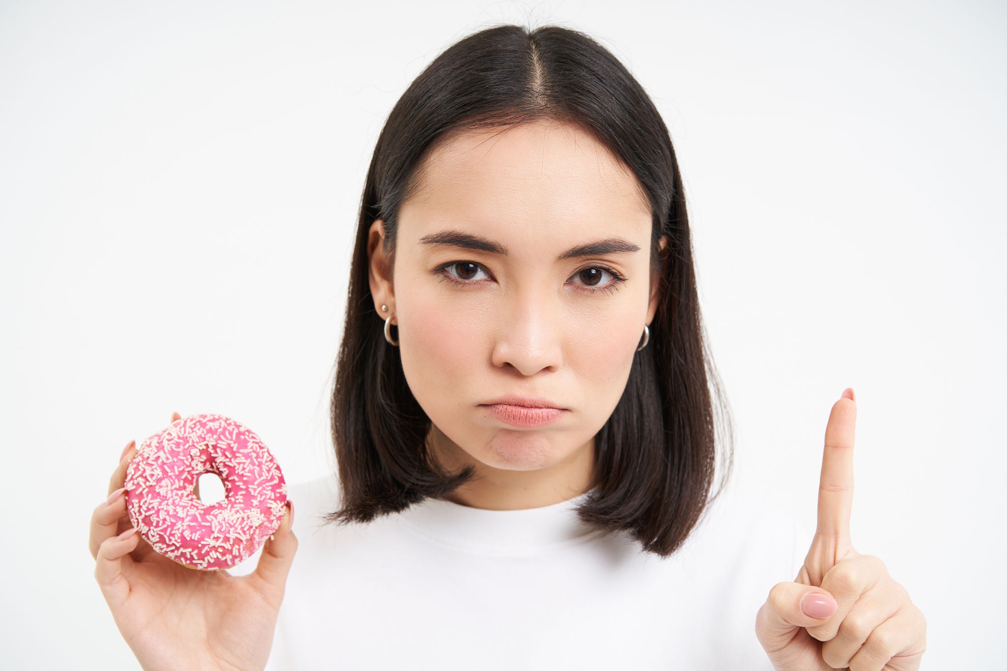 Close up of serious asian woman, shows taboo, stop gesture and pink glazed doughnut, forbids eating sweets, white background.