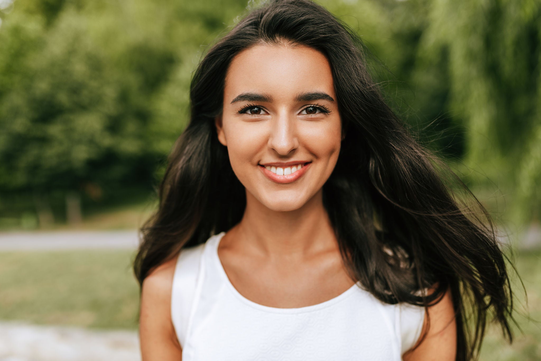 Outdoor close-up portrait of a beautiful young brunette woman smiling broadly and looking to the camera, posing against nature background in the park. Pretty female walking in the park. 